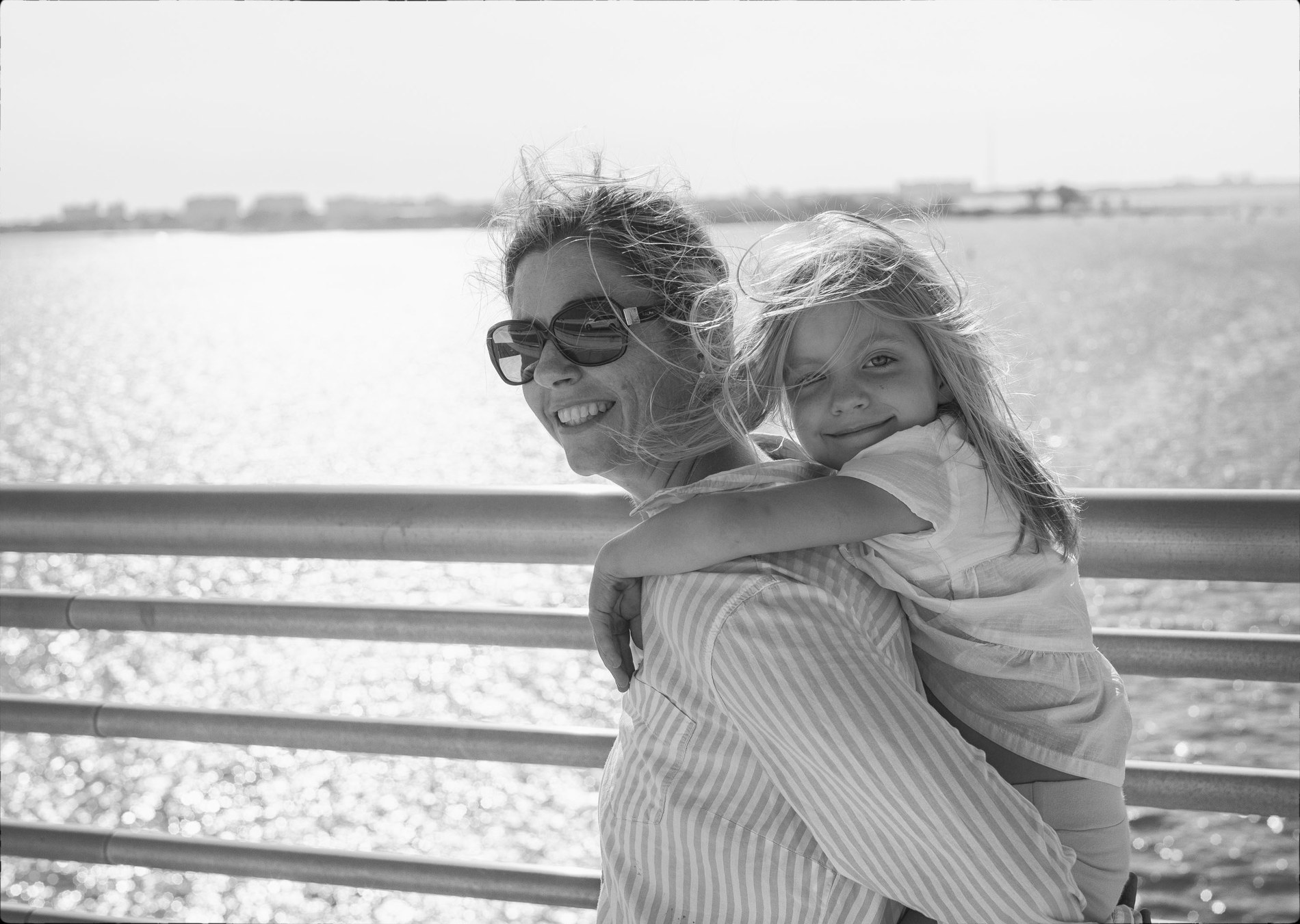 Julianne carrying Allie on her back on a waterfront bridge in Sarasota, both smiling into the camera, hair blown by the wind