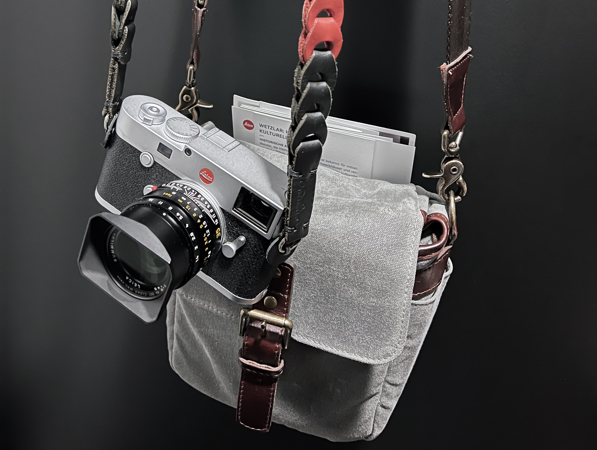 Silver Leica M rangefinder with a 35mm Summilux hanging on a worn gray canvas camera bag with braided leather straps