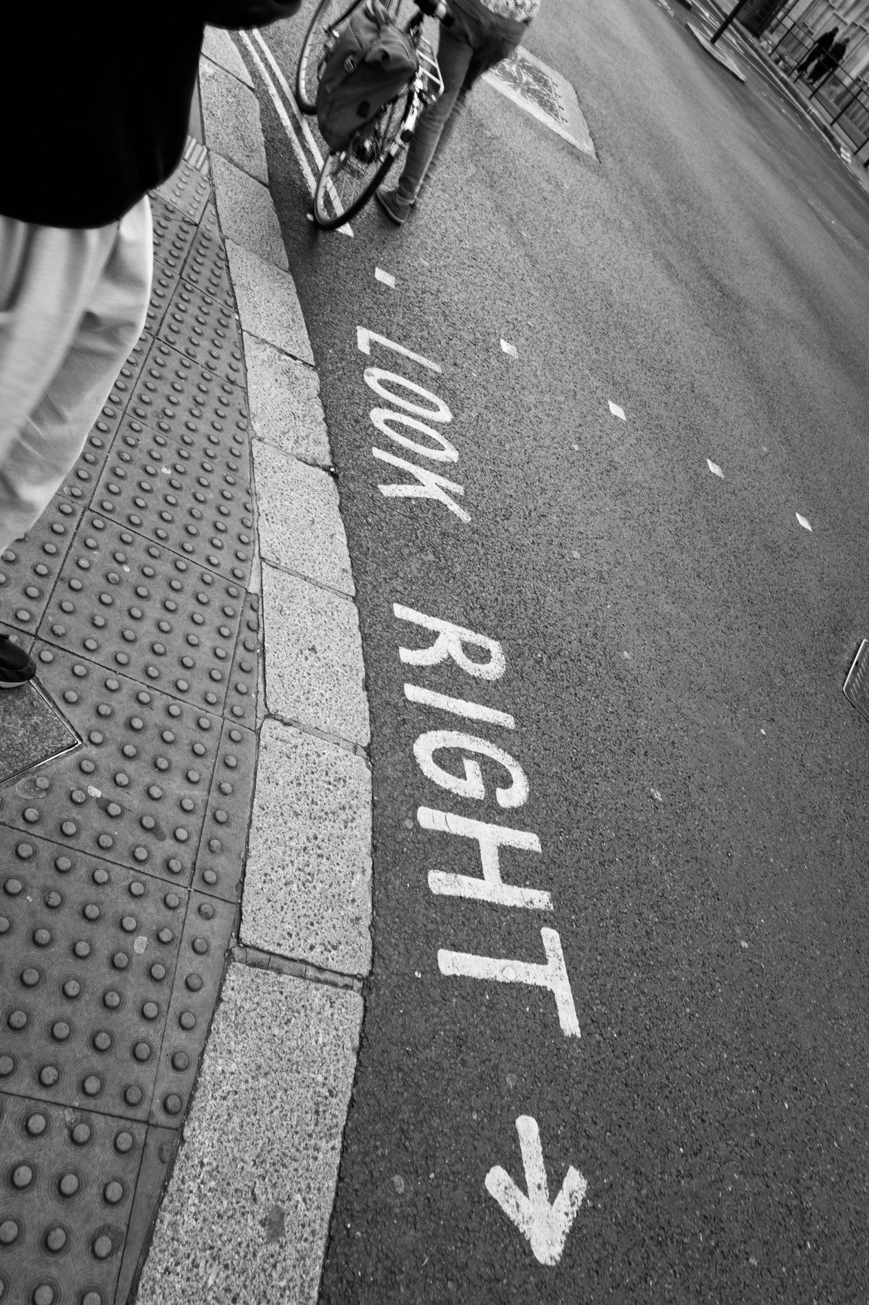 LOOK RIGHT painted on a London street beside tactile paving, a bicycle rolling past and a pedestrian at the edge of the frame