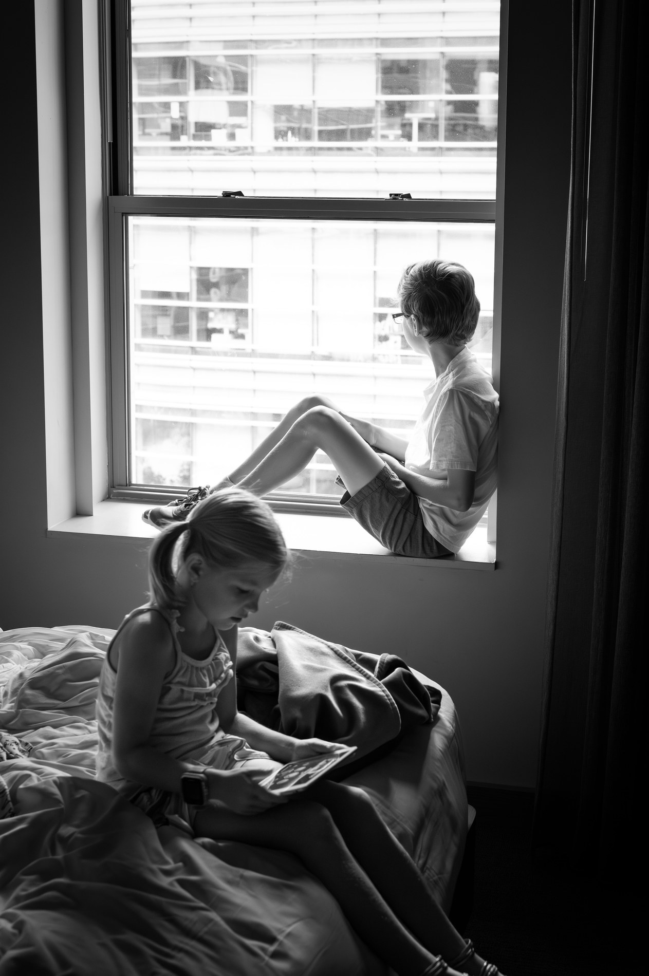 Hotel room in New York: Adam sits in a bright window looking out at the building opposite, Allie sits on the bed looking at a tablet