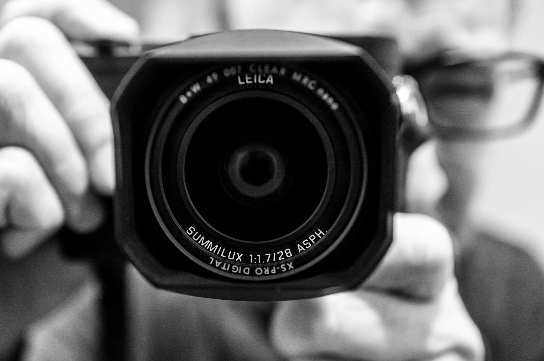 Close-up of a Leica Q-series 28mm Summilux f/1.7 lens looking straight into the camera, hands and glasses soft in the background